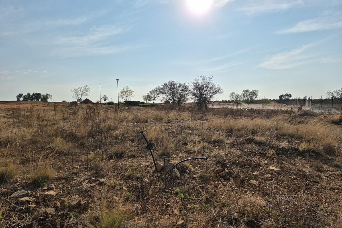 A view of a vacant residential plot featuring dry grass, scattered rocks, and visible utility connection points under a bright sky.