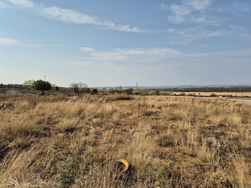 A wide-angle view of a vacant residential plot featuring dry natural vegetation, perimeter fencing, and distant landscape views.