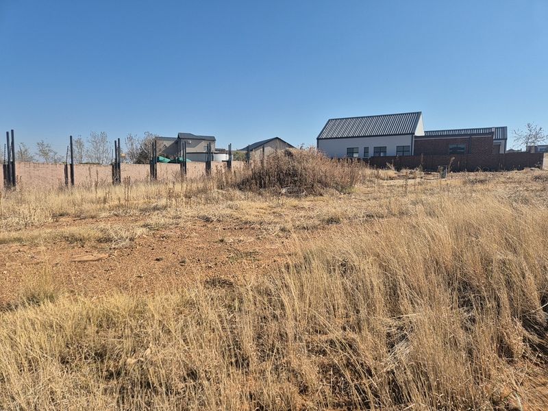 A vacant residential plot featuring dry grass, perimeter walling, and installed electric fencing, situated in a developed neighborhood.