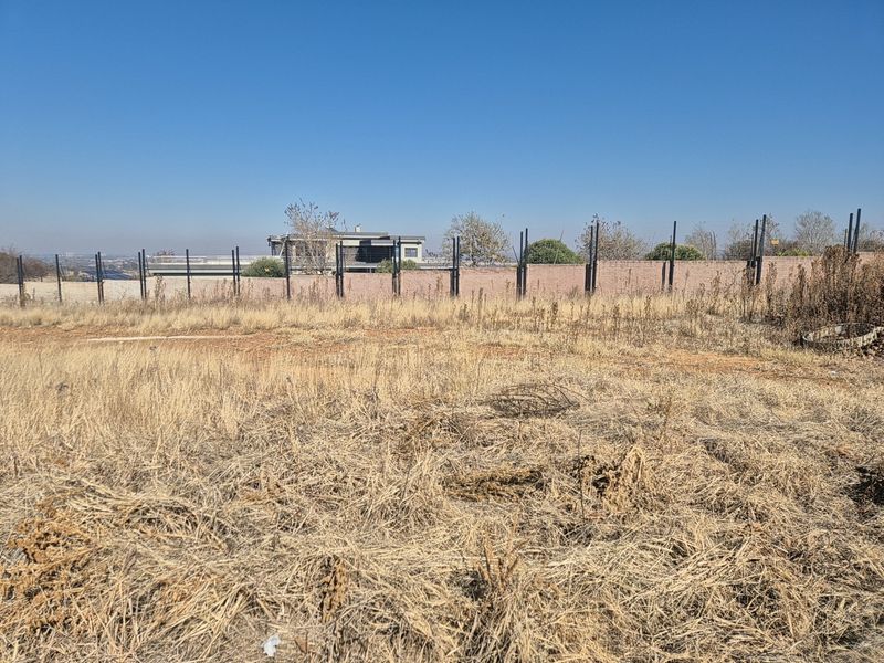 A view of a vacant residential plot featuring dry grass and a perimeter wall with metal fence posts.