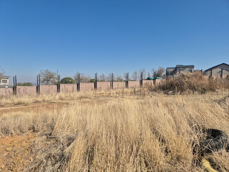 A vacant residential plot featuring dry grass, perimeter brick walling, and metal fence posts under a clear blue sky.