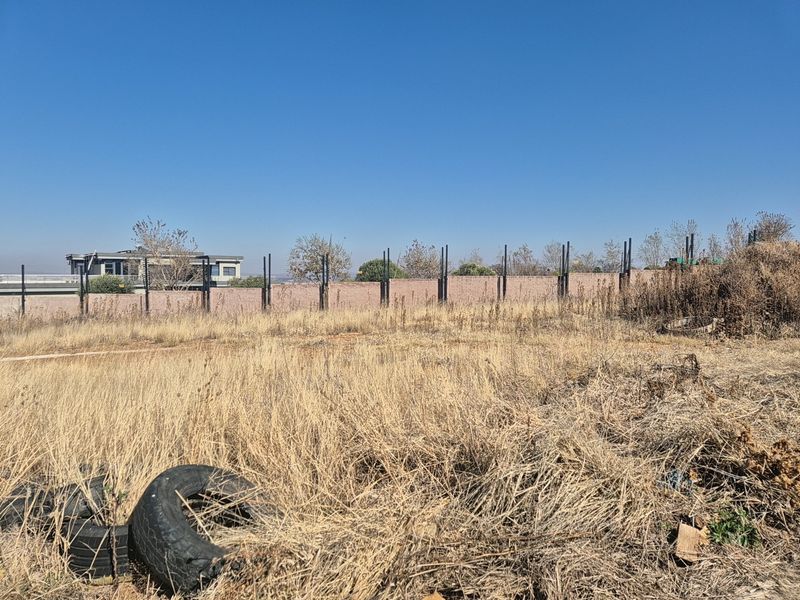 A vacant residential plot featuring dry, overgrown grass and a perimeter brick wall with metal fence posts.