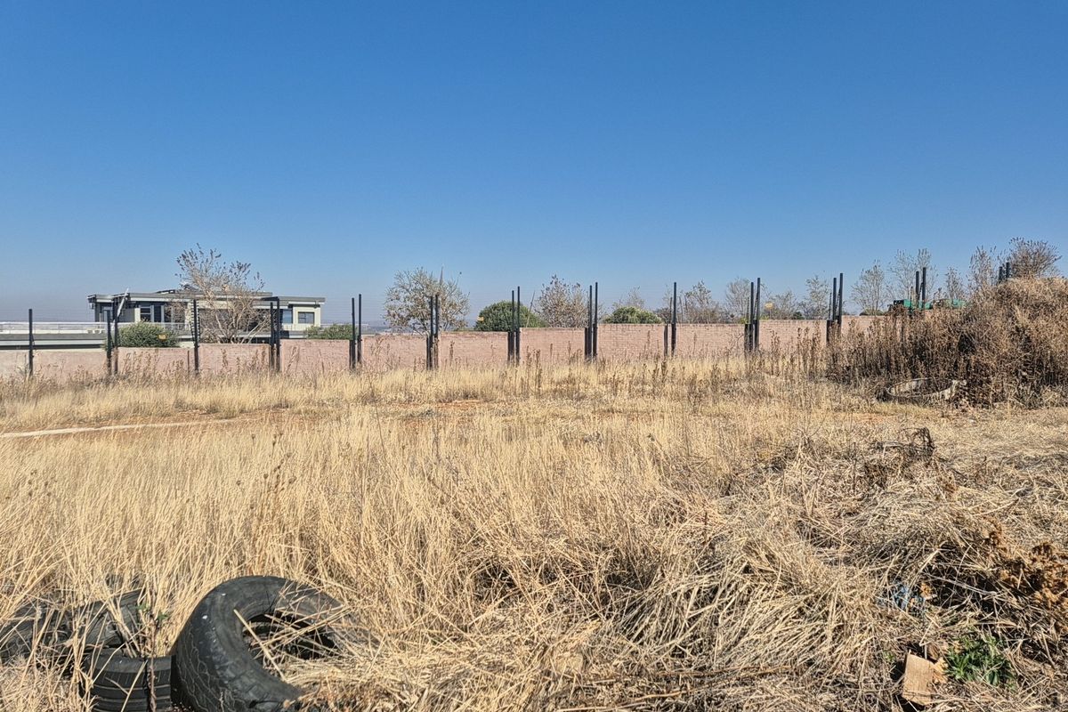 A vacant residential plot featuring dry, overgrown grass and a perimeter brick wall with metal fence posts.