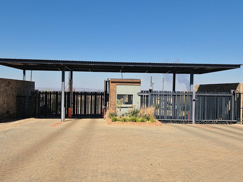 A secure residential estate entrance featuring a paved driveway, metal security gates, and a modern guard house with stone-clad perimeter walls.