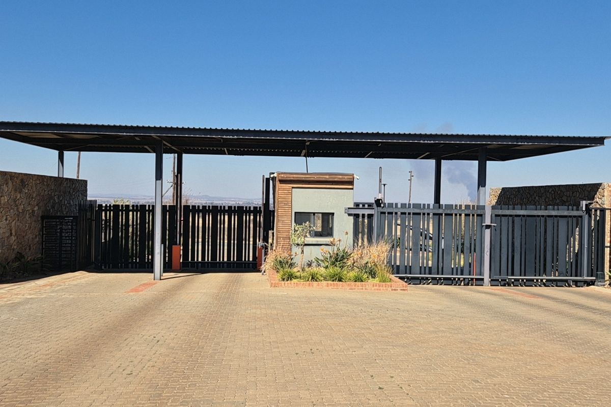 A secure residential estate entrance featuring a paved driveway, metal security gates, and a modern guard house with stone-clad perimeter walls.