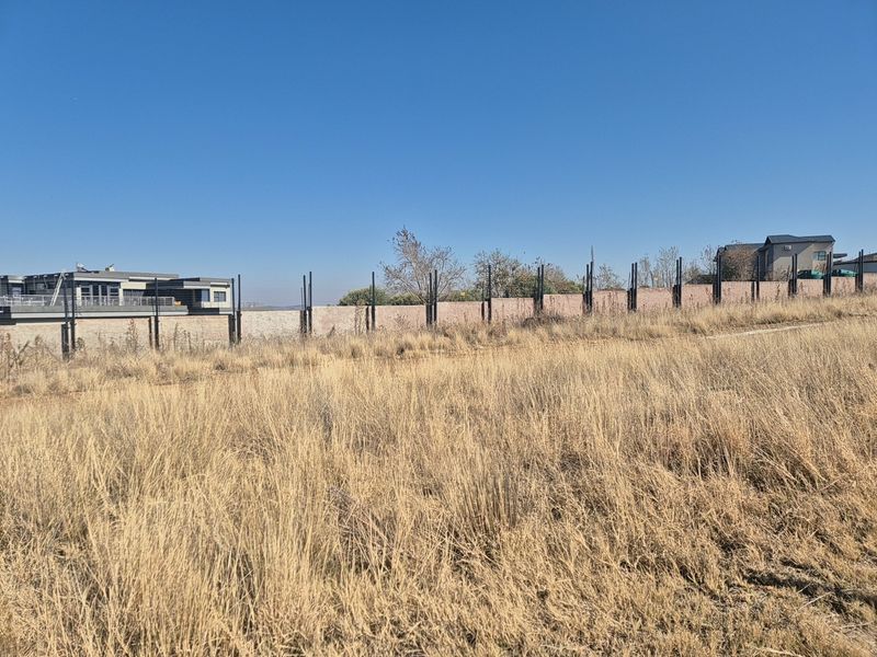 A view of a vacant residential plot featuring dry grass and a perimeter wall with metal fence posts.