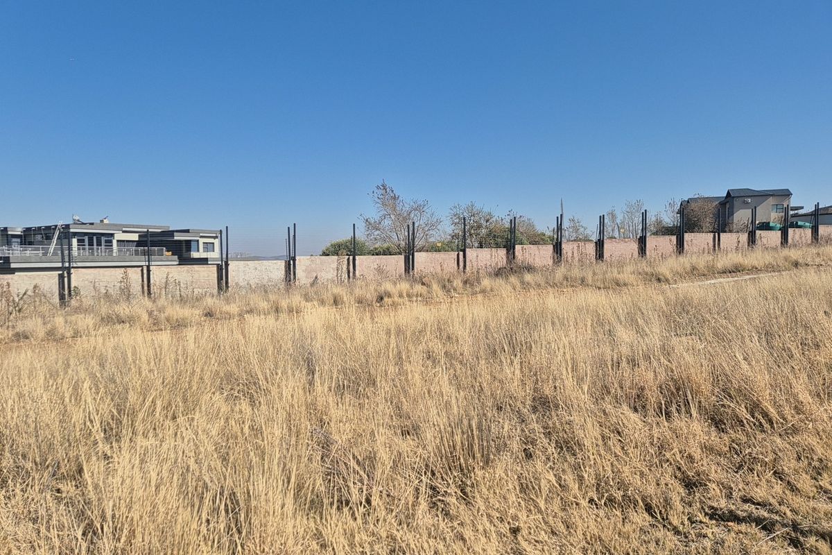 A view of a vacant residential plot featuring dry grass and a perimeter wall with metal fence posts.
