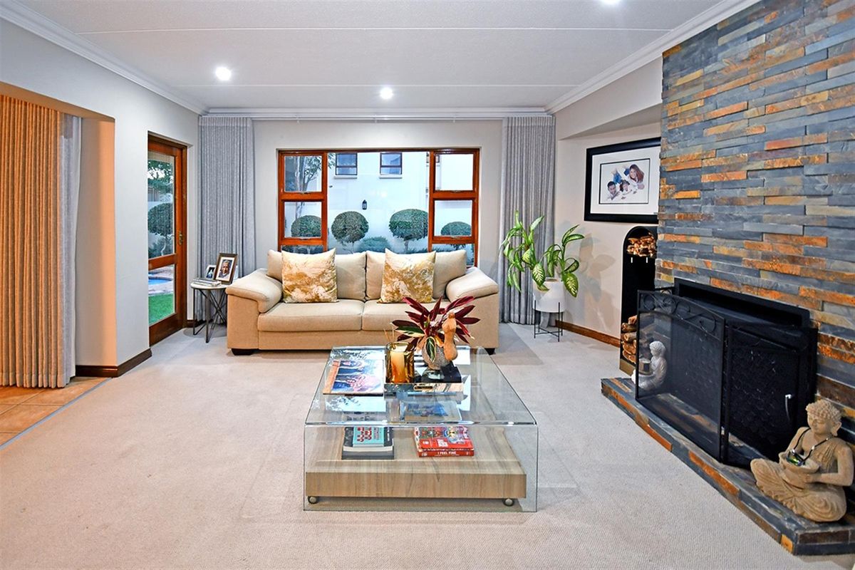 A comfortable living room featuring a prominent stone-clad fireplace, neutral carpeting, and large windows providing natural light and garden views.