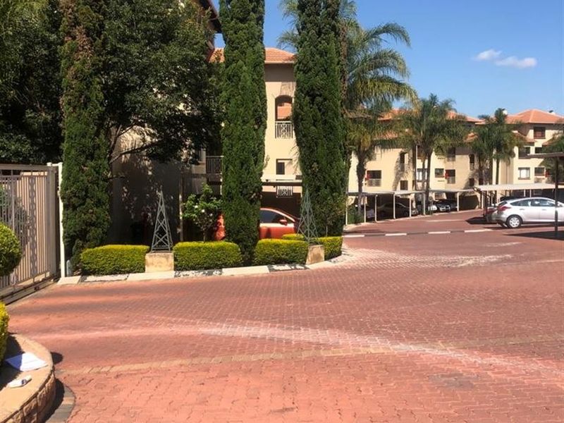 View of the apartment complex entrance featuring a paved driveway, security gate, and established landscaping with covered parking areas.