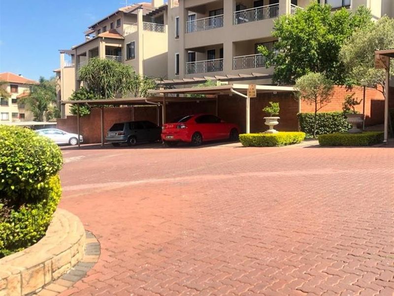 Exterior view of a multi-story apartment complex featuring covered parking bays, paved driveway, and private balconies with well-maintained landscaping.