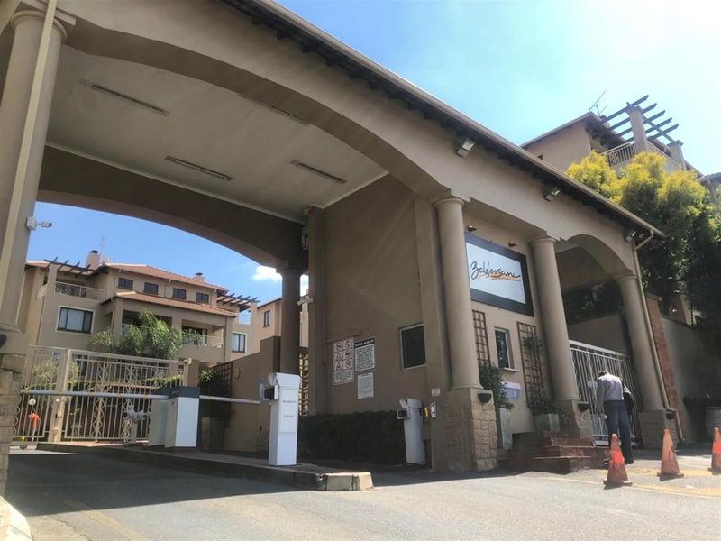 The main entrance and security gatehouse of the apartment complex, featuring a large architectural archway and controlled access boom gates.