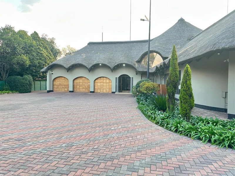 Front elevation of a property featuring a distinctive thatched roof, triple garage doors, and a spacious paved driveway with established garden beds.