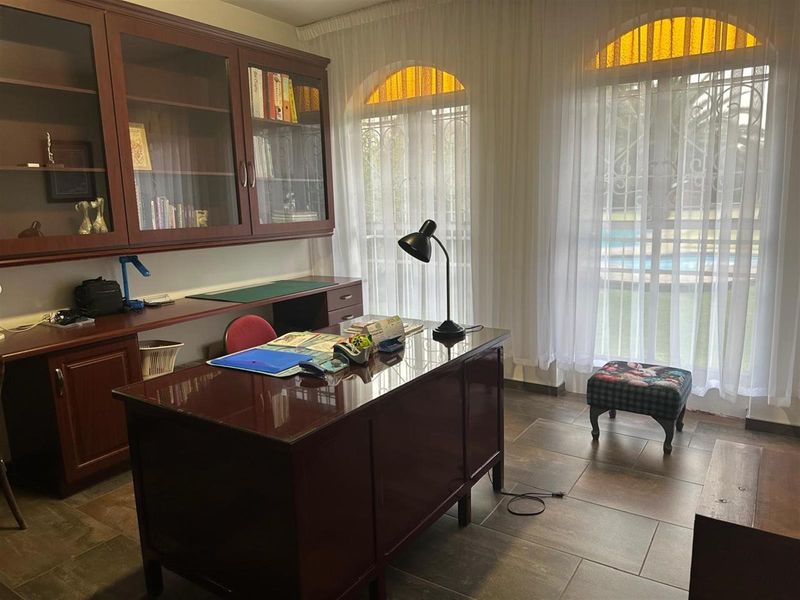 A home office featuring built-in wooden cabinetry, tiled flooring, and large arched windows with security bars overlooking the garden.