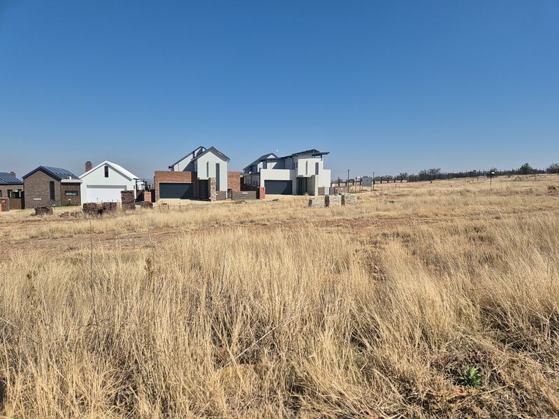 A view of a vacant residential plot featuring dry grass, with established modern homes visible in the background.
