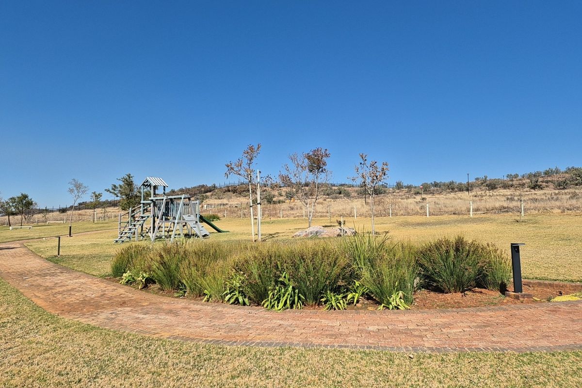 A well-maintained residential garden featuring a paved walkway, landscaped garden beds, and a children's playground set within an open green space.