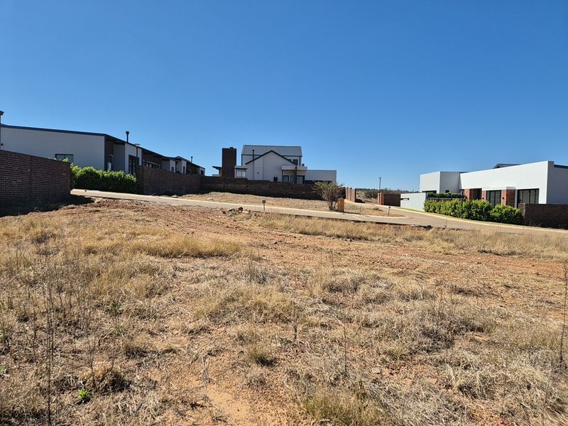 A vacant residential plot featuring natural grass terrain, adjacent to a paved road and established modern residential properties in a secure estate.