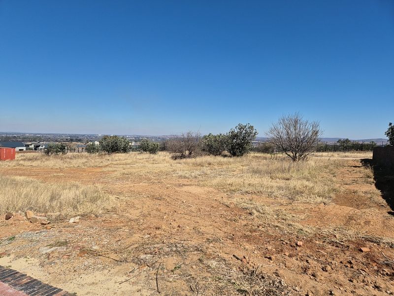 A wide-angle view of a vacant residential plot featuring dry grass, native shrubs, and a distant horizon under a clear blue sky.