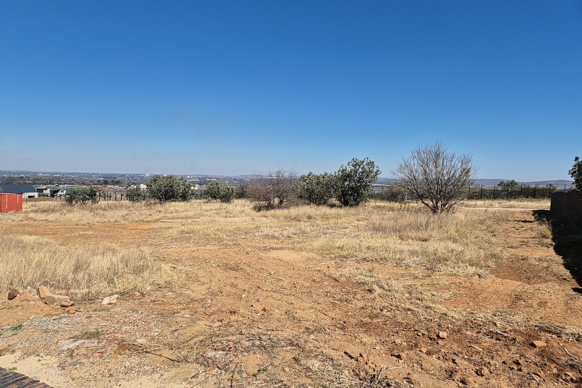 A wide-angle view of a vacant residential plot featuring dry grass, native shrubs, and a distant horizon under a clear blue sky.