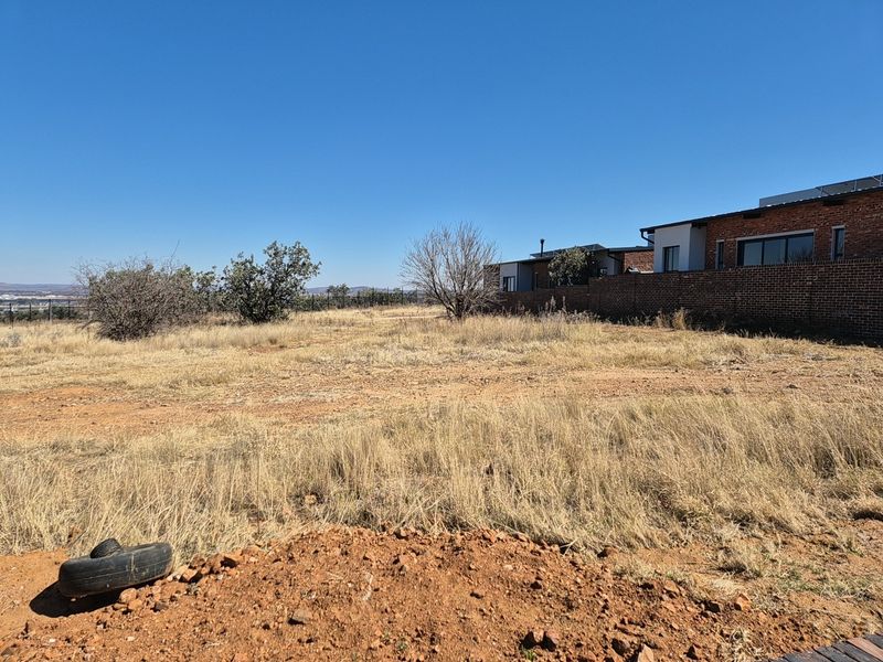 A vacant residential plot featuring dry grass, natural terrain, and an existing brick boundary wall adjacent to neighboring properties.
