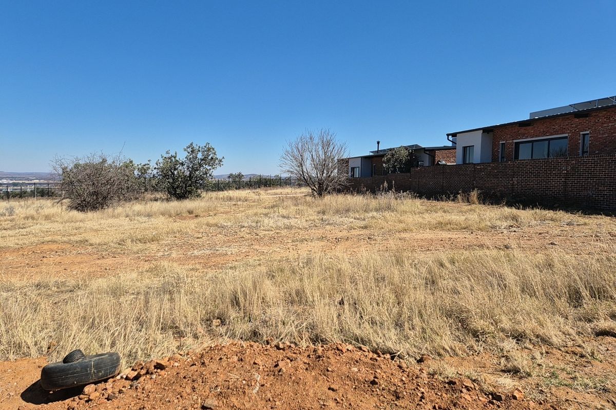 A vacant residential plot featuring dry grass, natural terrain, and an existing brick boundary wall adjacent to neighboring properties.