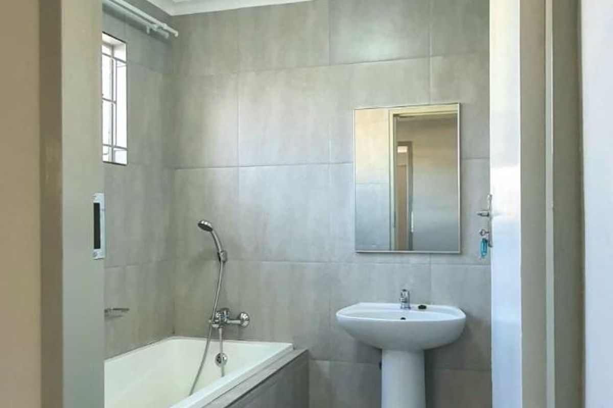 A clean, modern bathroom featuring grey wall and floor tiles, a white pedestal basin, and a built-in bathtub with a handheld shower fixture.
