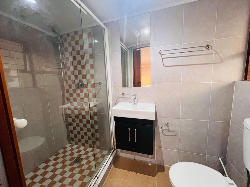 A functional bathroom featuring a glass-enclosed shower, wall-mounted vanity with basin, and neutral wall tiling with a decorative checkered accent.