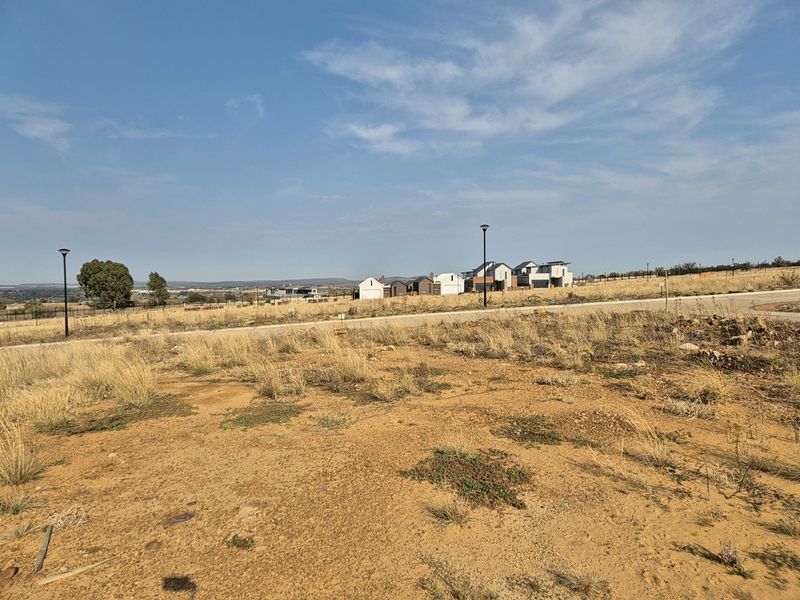 A view of a vacant residential plot featuring a paved road and street lighting, with neighboring houses visible in the distance.