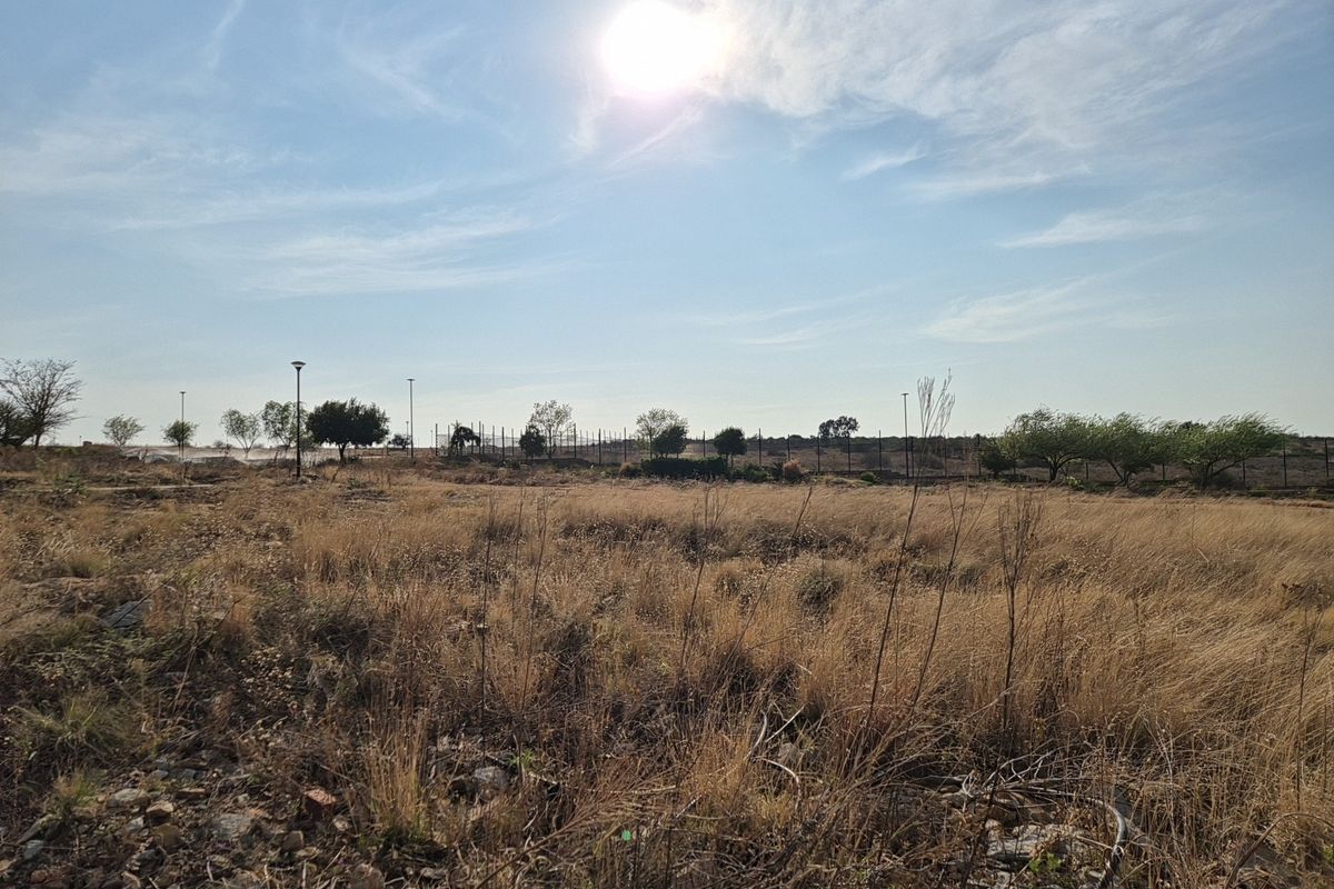 A view of a vacant residential plot featuring dry grass, perimeter fencing, and street lighting under a clear sky.