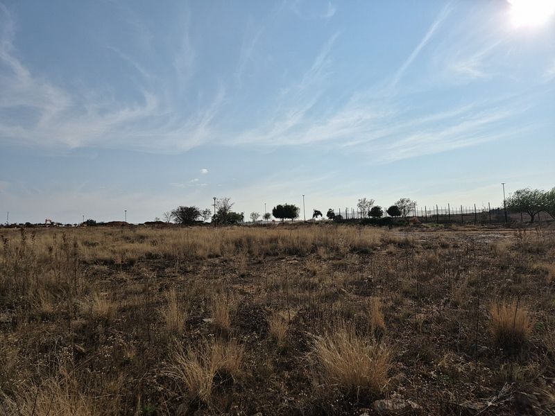 A view of a vacant residential plot featuring dry grass, perimeter fencing, and distant street lighting infrastructure.