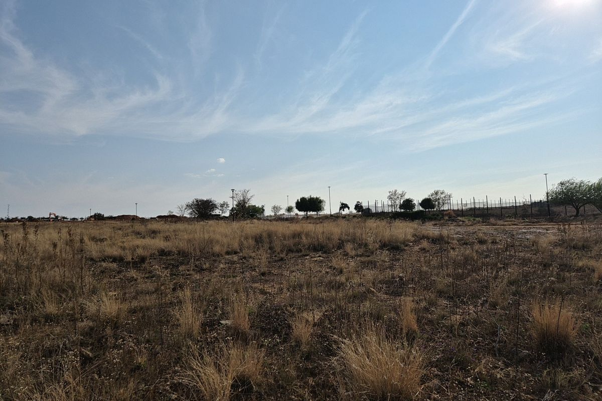 A view of a vacant residential plot featuring dry grass, perimeter fencing, and distant street lighting infrastructure.