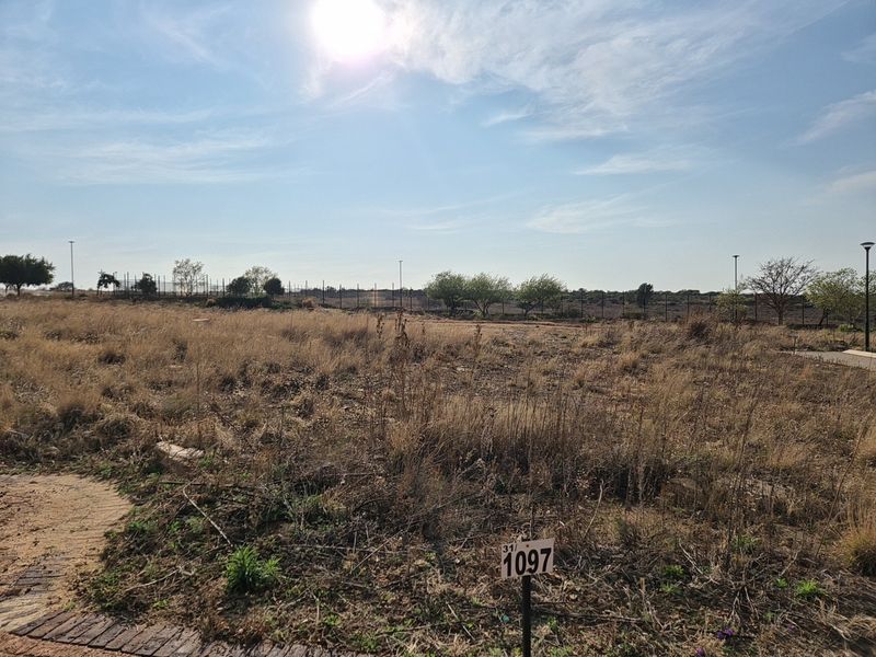 A vacant residential plot featuring dry grass, perimeter fencing, and street lighting, ready for development.