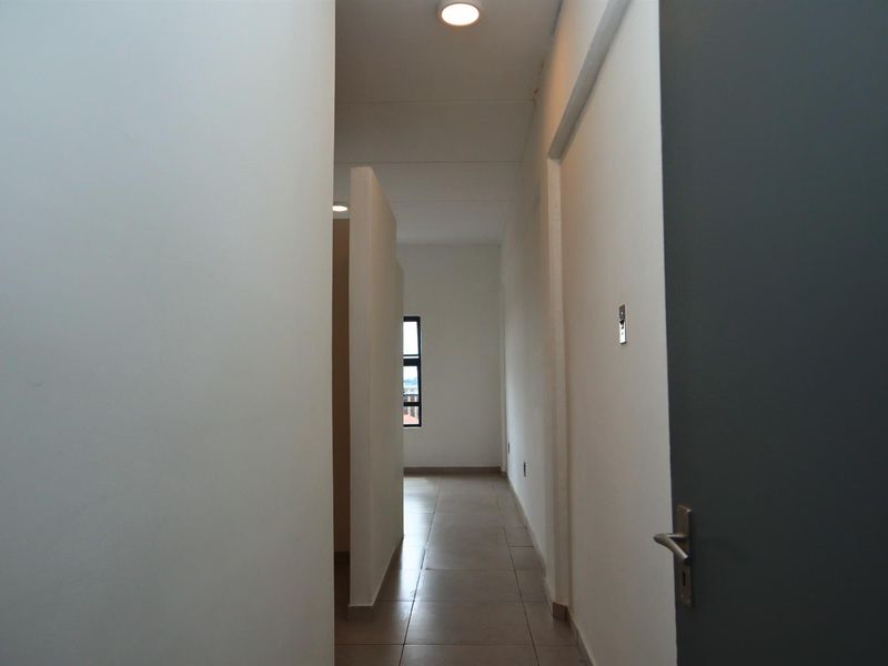 A clean, minimalist hallway featuring neutral tiled flooring, recessed ceiling lighting, and a clear view through to an adjacent room with a window.