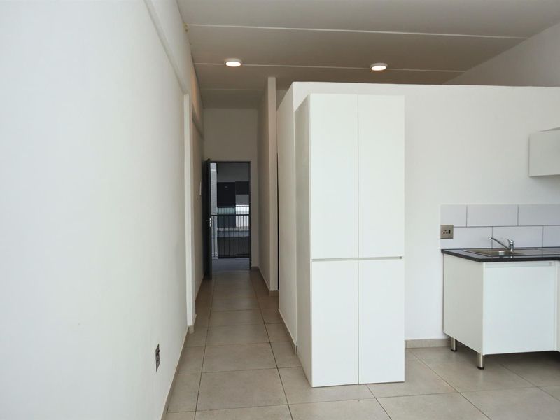 An open-plan living and kitchen area featuring tiled flooring, built-in white cabinetry, and a functional sink, leading into a hallway.