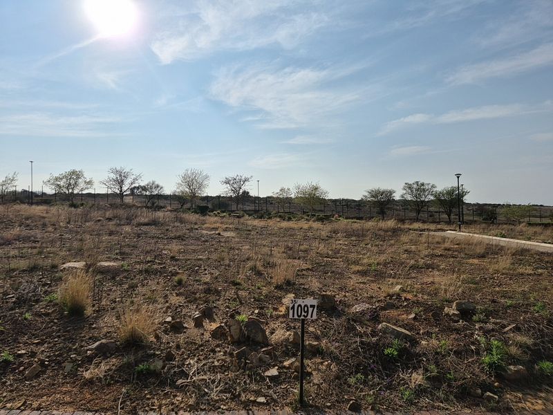 A vacant residential plot featuring natural terrain, perimeter fencing, and street lighting, ready for development.