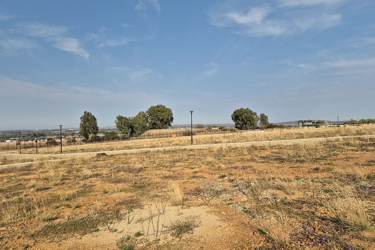 A vacant residential plot featuring natural terrain, paved road access, and established perimeter fencing in a developing estate setting.