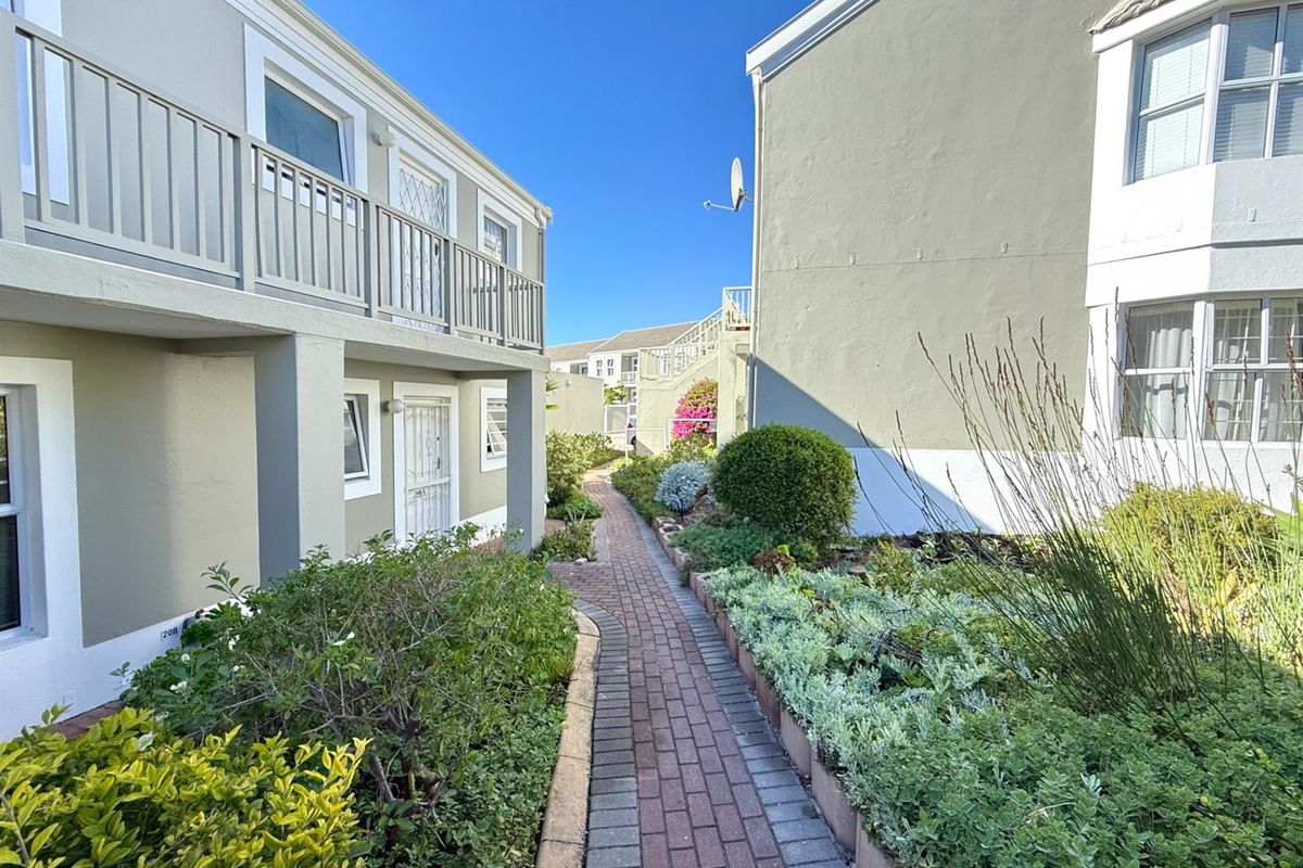 A well-maintained exterior walkway between apartment buildings, featuring paved pathways, landscaped garden beds, and access to ground-floor units and upper-level balconies.