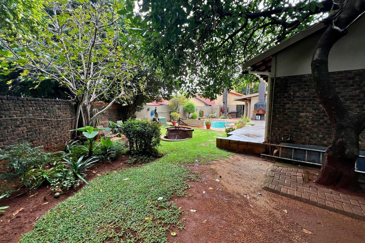 A spacious rear garden featuring a lawn area, mature trees, brick boundary walls, and a view towards the swimming pool and outdoor entertainment area.
