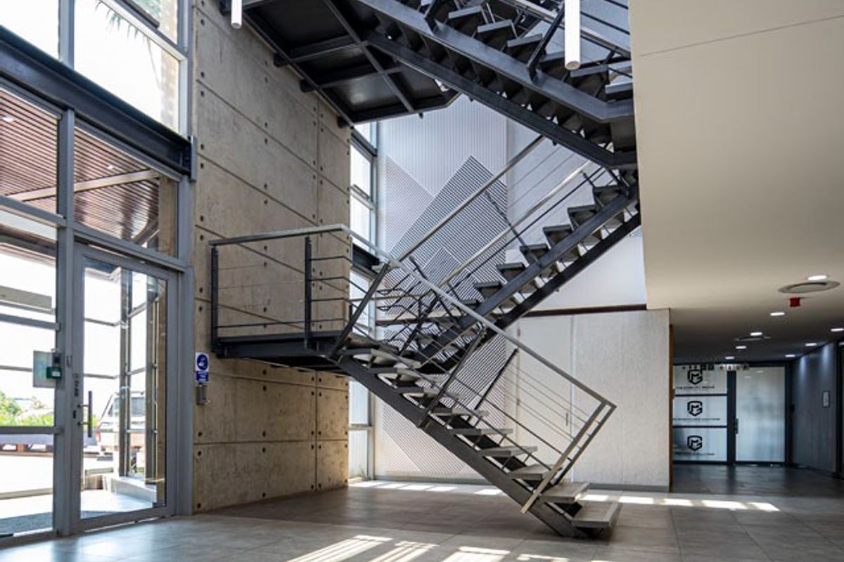 Modern commercial lobby featuring a striking industrial-style steel staircase, exposed concrete wall, large glass facade, and sleek tiled flooring.