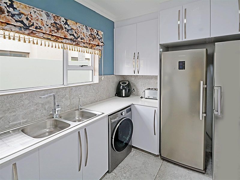 A modern, functional kitchen featuring white cabinetry, stone-look countertops, a double stainless steel sink, and integrated space for laundry appliances.