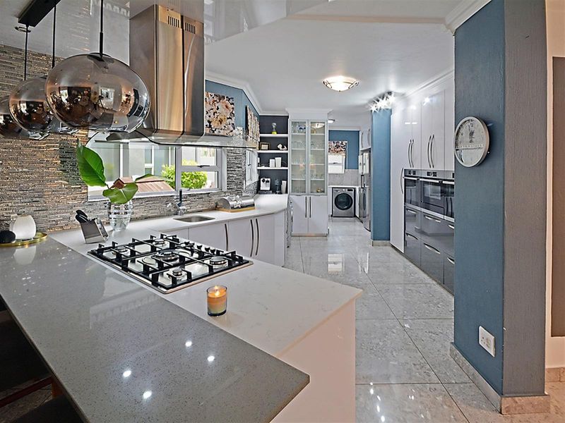 Modern open-plan kitchen featuring stone countertops, integrated gas hob, stainless steel extractor fan, built-in ovens, and high-gloss cabinetry with tiled flooring.