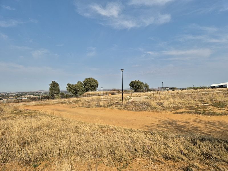 A view of a vacant residential plot featuring an unpaved access road, perimeter fencing, and installed street lighting under a clear sky.
