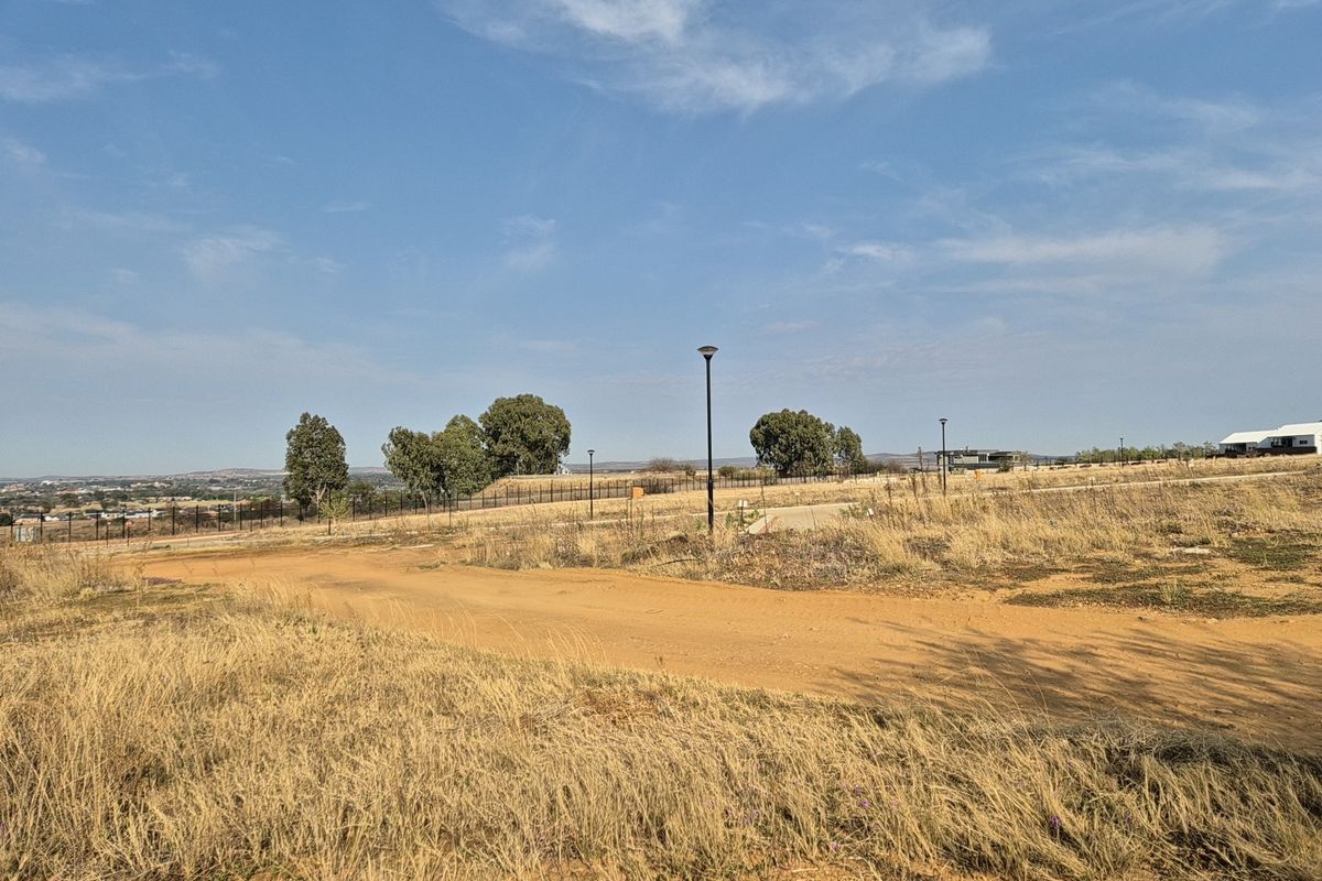 A view of a vacant residential plot featuring an unpaved access road, perimeter fencing, and installed street lighting under a clear sky.
