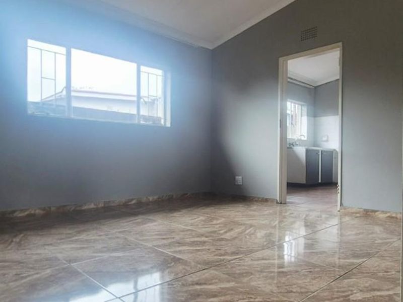A bright, empty living area featuring polished marble-look floor tiles, neutral grey walls, and a doorway leading to an adjacent kitchen space.