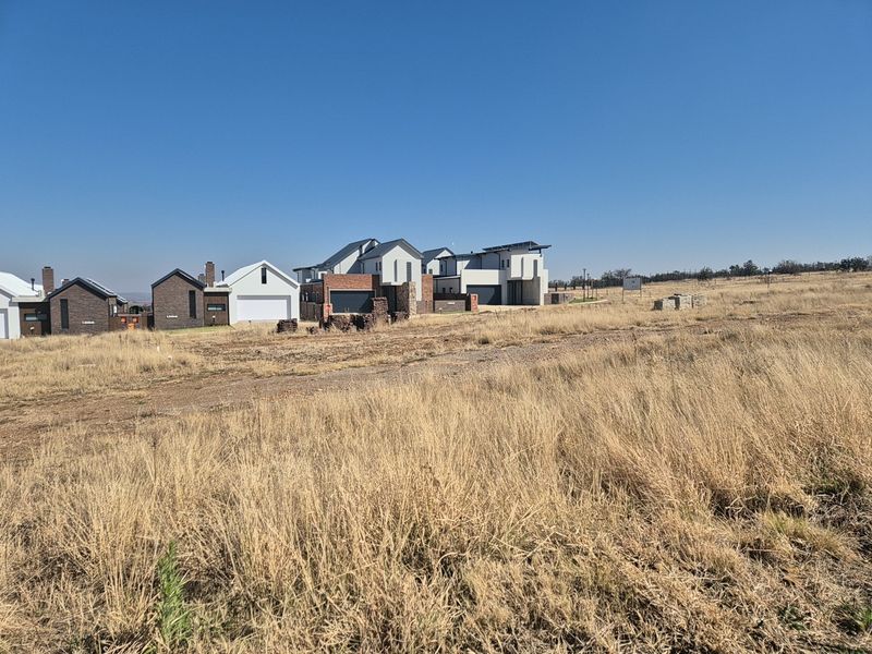 A view of a vacant residential plot situated within a developing estate, featuring surrounding modern homes and open grass terrain.