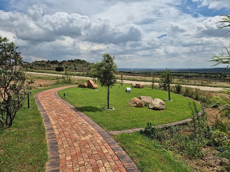 A well-maintained residential garden featuring a brick paved pathway, manicured lawn, and decorative boulders with scenic landscape views.