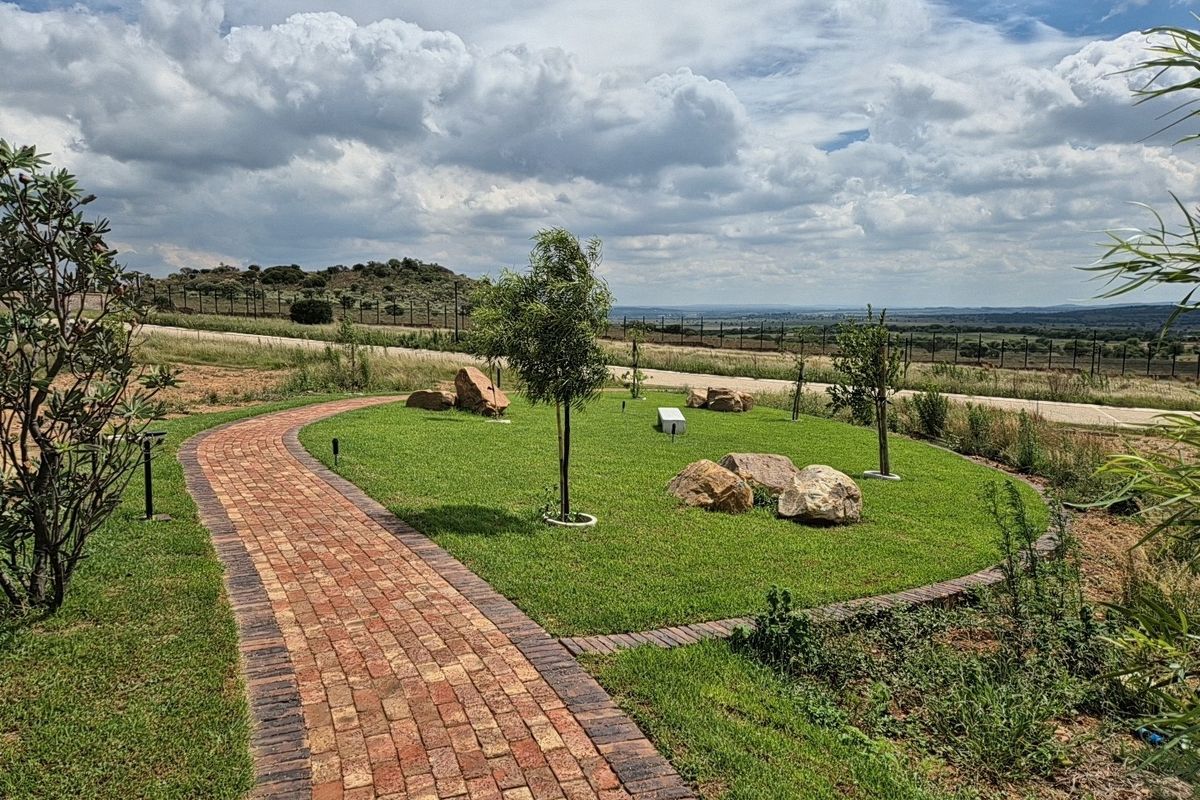 A well-maintained residential garden featuring a brick paved pathway, manicured lawn, and decorative boulders with scenic landscape views.