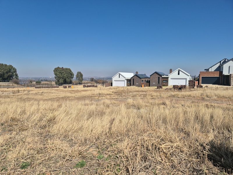 A view of a vacant residential plot with dry grass, showing neighboring modern homes in the background.