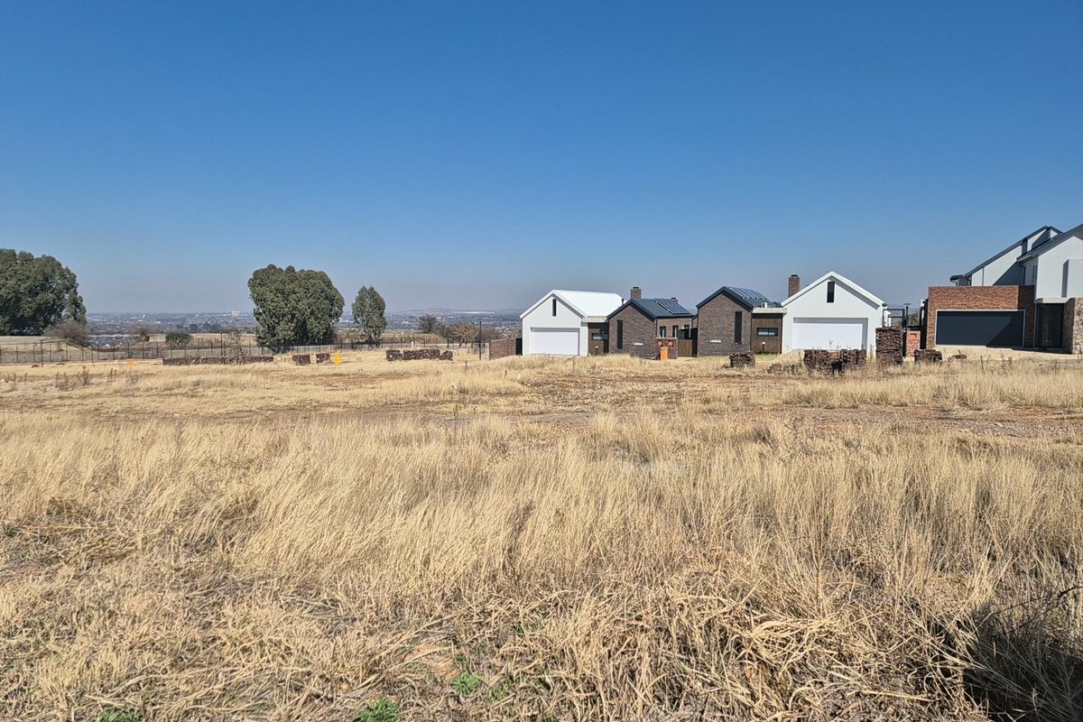 A view of a vacant residential plot with dry grass, showing neighboring modern homes in the background.