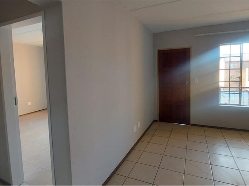 An empty, neutrally painted living area featuring tiled flooring, a timber door, and a window with security bars, offering a clean blank canvas.