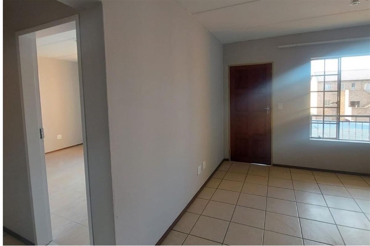 An empty, neutrally painted living area featuring tiled flooring, a timber door, and a window with security bars, offering a clean blank canvas.